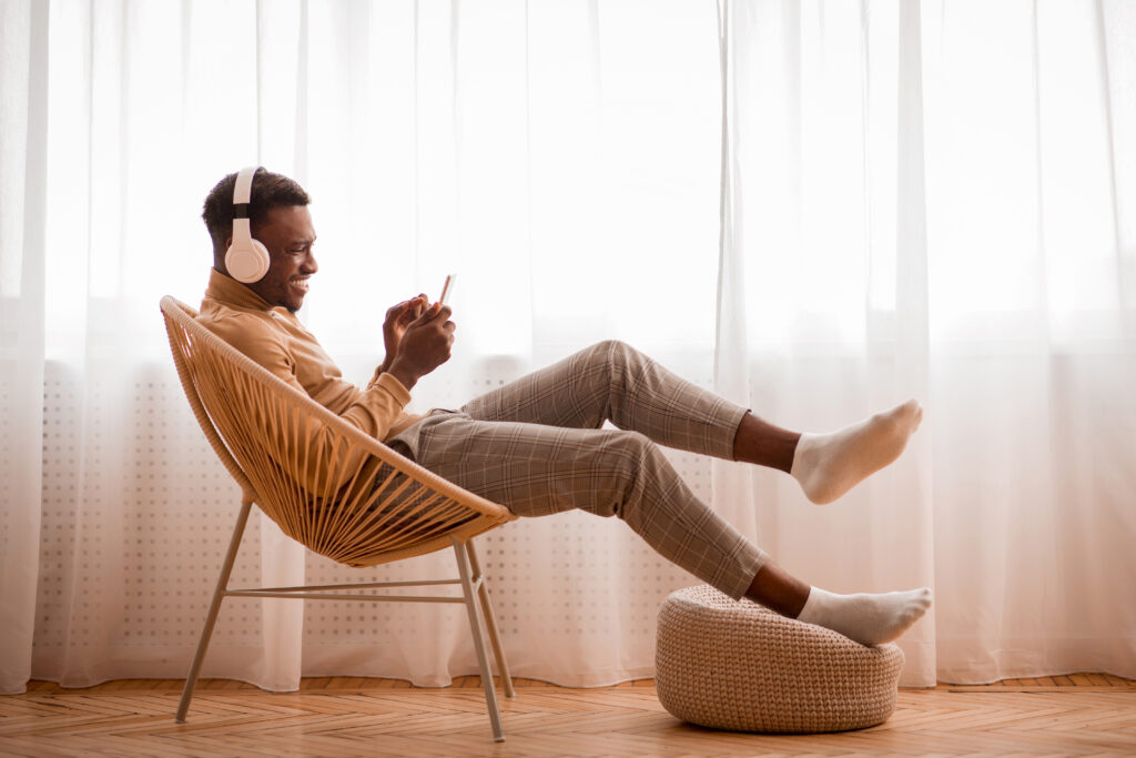 RootsBNB Guy In Headphones Using Smartphone Sitting On Chair Indoor.